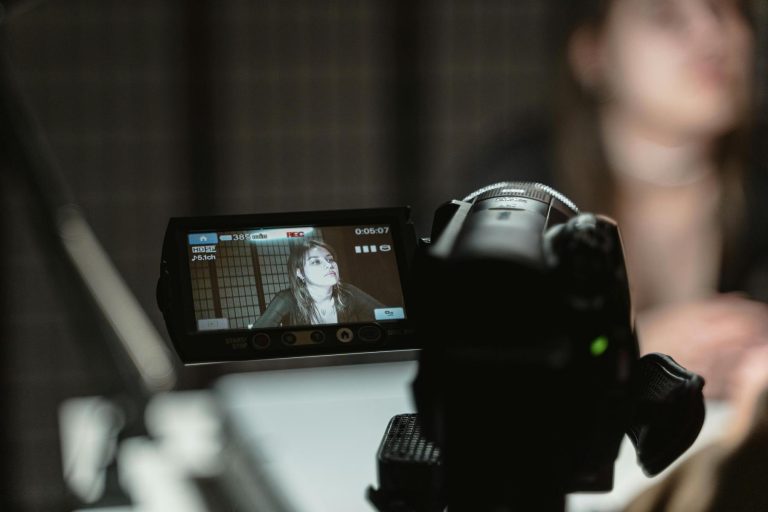A camera records a woman sitting at a table; her image is visible on the camera’s display screen while she is out of focus in the background.