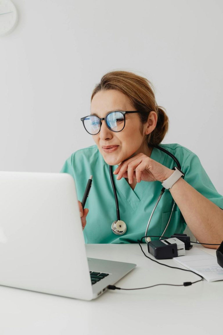 A healthcare professional in green scrubs with a stethoscope sits at a desk, looking at a laptop and holding a pen, with medical equipment and papers nearby.