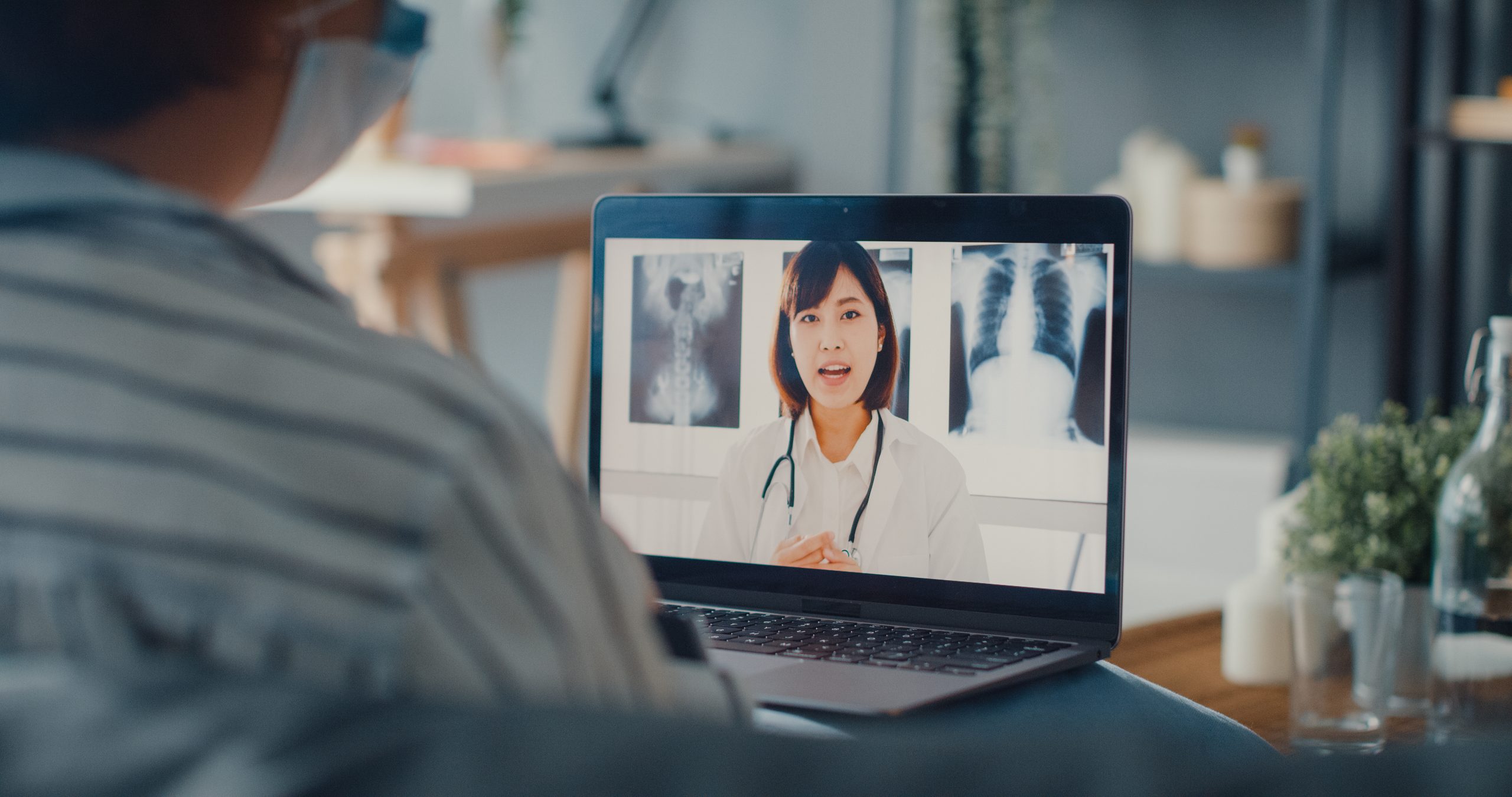 A person wearing a mask participates in a video call with a doctor, who is visible on a laptop screen with medical images in the background.