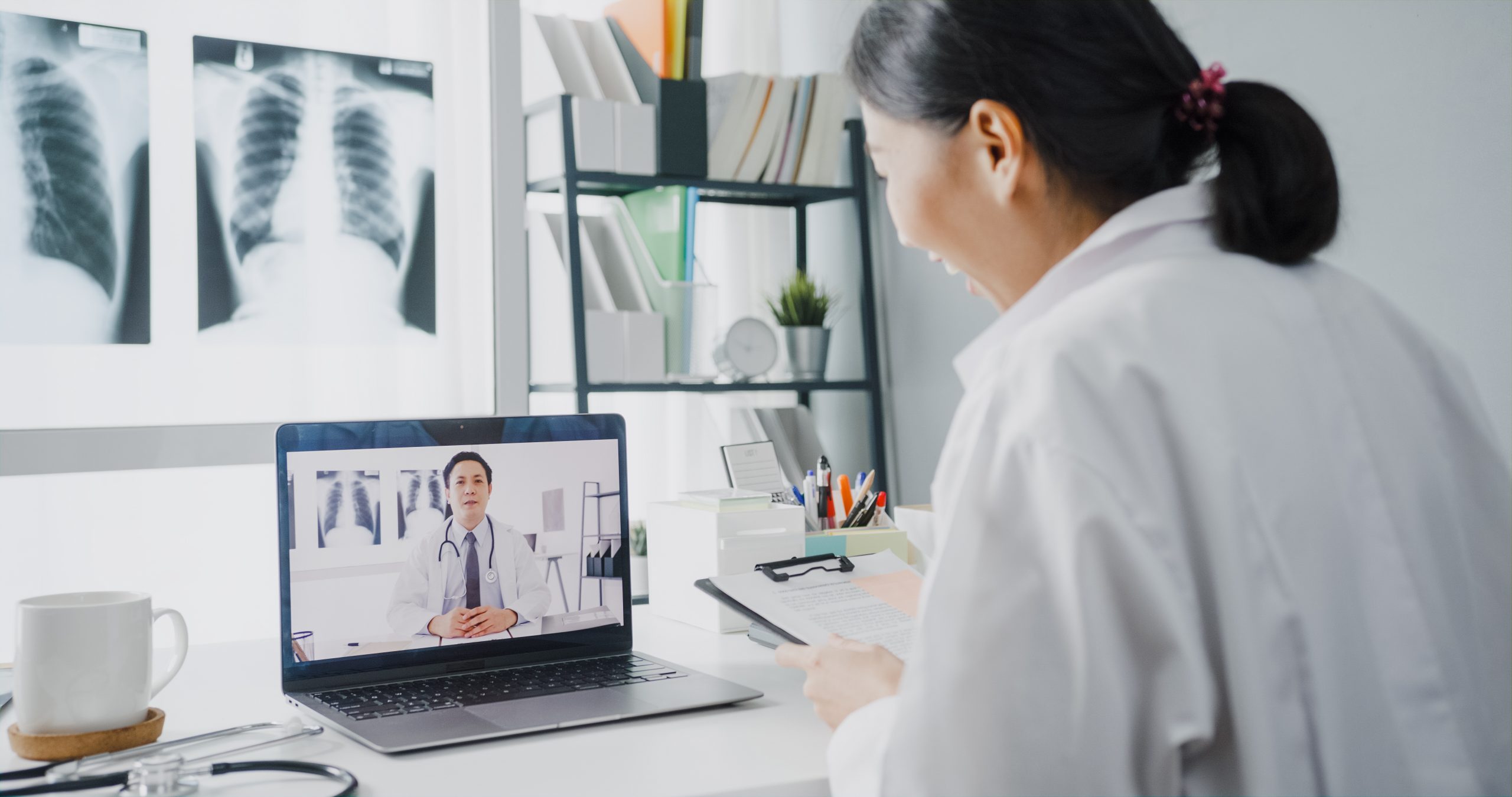 A doctor sits at a desk holding a clipboard and has a video call with another doctor on a laptop screen; chest X-rays are visible in the background.