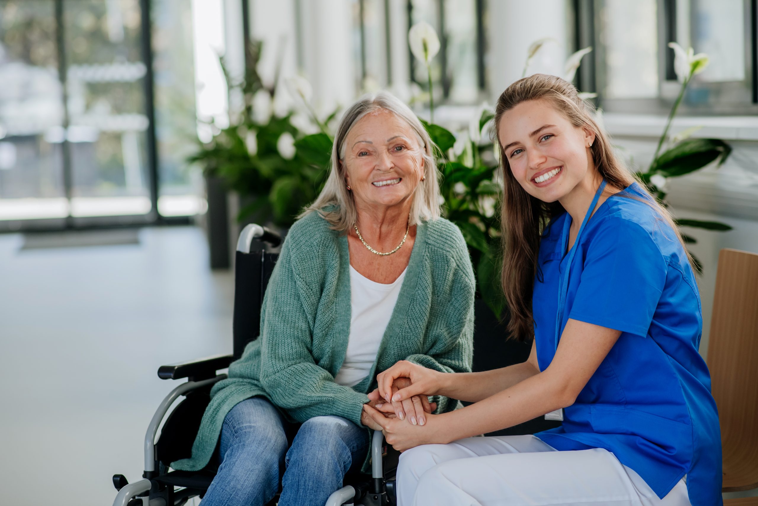 An older woman in a wheelchair sits next to a smiling caregiver in blue scrubs. They are holding hands in a bright, indoor setting with green plants in the background.